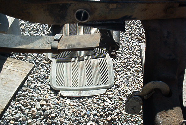 Ribbed metal step plate under vehicle frame with leaf spring and tow hook, over gravel