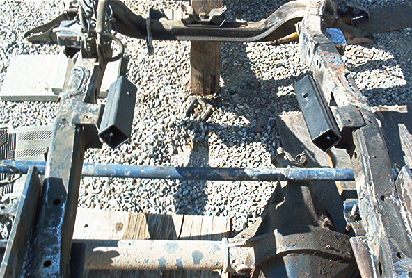 Bare truck frame on gravel with black body mount brackets and rear axle visible during frame-off restoration
