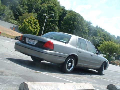 2000 Ford Crown Victoria - A silver 2000 Ford Crown Victoria is parked at an angle in a lot, with trees and a hill in the background.