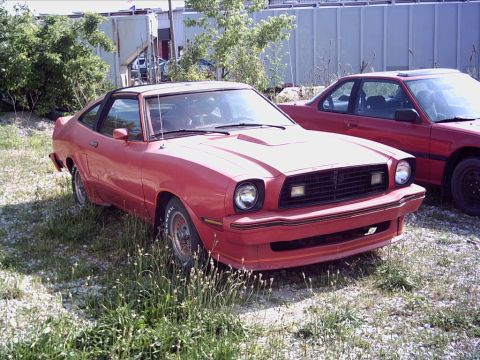 1900 Ford Wagon - A red coupe with a hood scoop is parked next to another red car in an outdoor lot.
