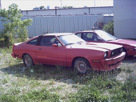 1900 Ford Wagon - A red 1978 Ford Mustang II King Cobra is parked in a grassy lot next to another vehicle.