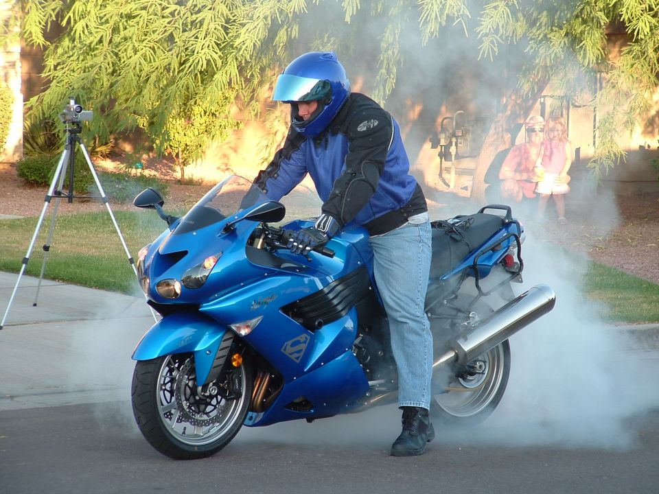 Blue motorcycle with a rider doing a burnout, emitting smoke. A tripod with a camera is visible to the left