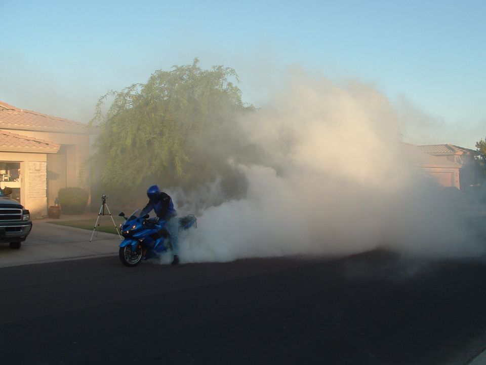 Blue motorcycle with rider in blue jacket and helmet doing a burnout, creating a large cloud of smoke. A pickup truck is parked to the left