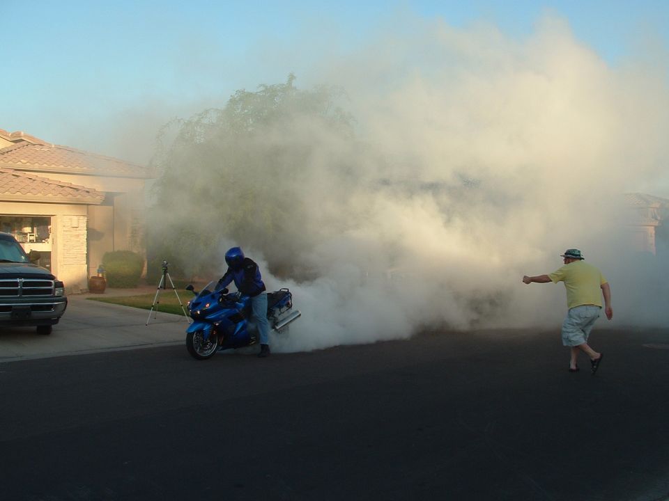 guy on blue motorcycle doing a burnout in a suburban street, another guy watches