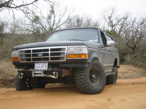 1995 Ford Bronco - This lifted Ford Bronco is parked on a dirt road with trees in the background.