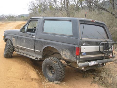 1995 Ford Bronco - A dirty black Ford Bronco with off-road tires is parked on a dirt road, showing its rear-left side.
