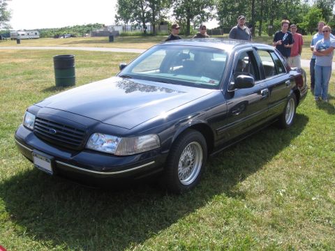 2000 Ford Crown Victoria - A dark blue 2000 Ford Crown Victoria sedan is parked on a grassy field with people in the background.