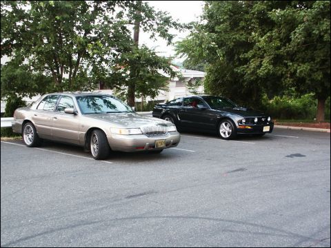 2001 Mercury Grand Marquis - Two cars, a Mercury Grand Marquis and a Ford Mustang, are parked in a lot with trees in the background.
