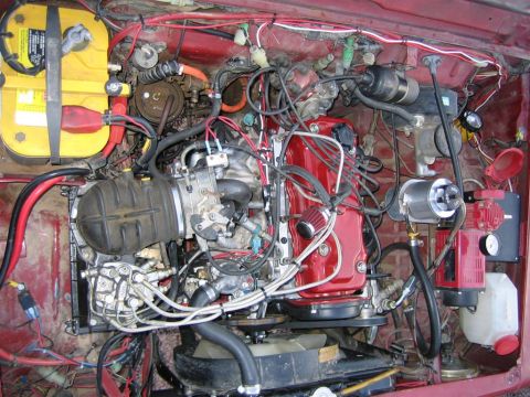 1987 Suzuki Samurai - Close-up view of a complex engine bay a red valve cover and a yellow Optima battery.