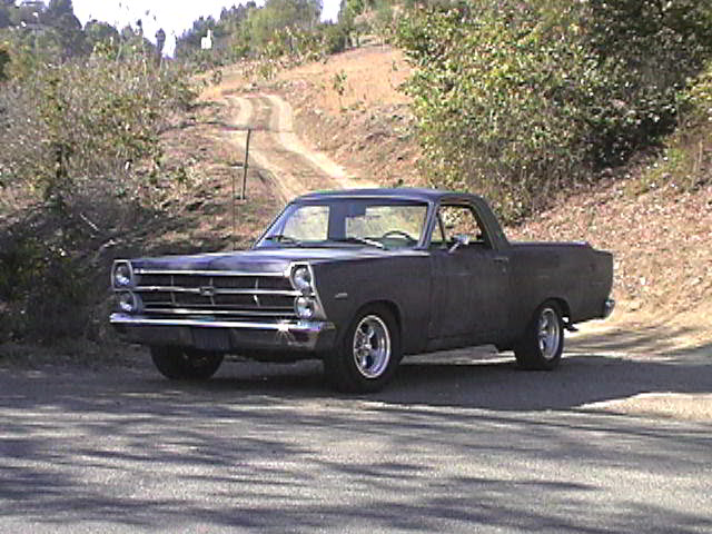Dark gray 1967 Ford Ranchero parked on pavement in front of a dirt hillside road, front three-quarter view