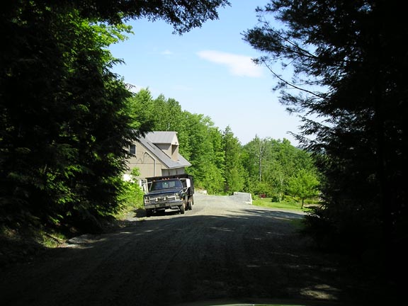Dark pickup truck parked on a gravel driveway in front of a house, surrounded by trees