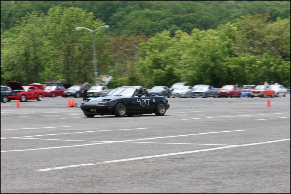 Black convertible sports car with "420 X" on the side, driving in a parking lot with other cars and orange cones