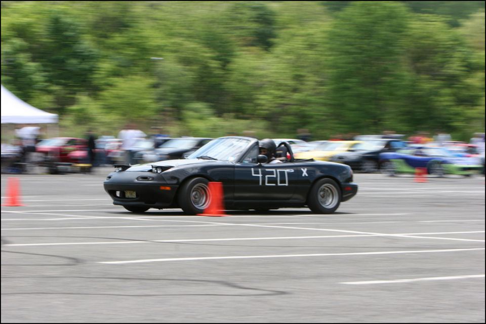 Black Mazda Miata convertible with racing numbers 420 X on the door, driving on a track with orange cones. Other cars are blurred in the background