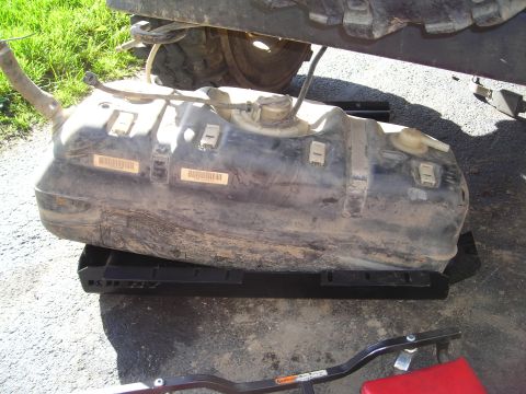 2003 Jeep TJ - A dirty fuel tank is shown removed from a vehicle, resting on a black mounting bracket. Two barcode labels are visible on the tank.