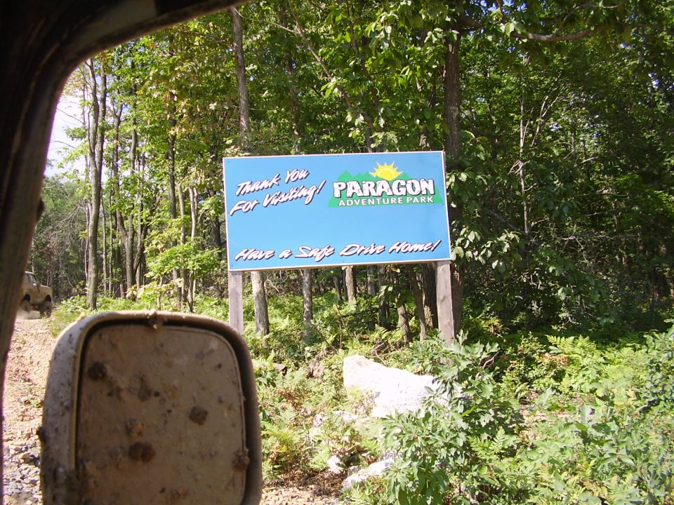 Sign at Paragon Adventure Park entrance. A dirt road leads into woods. Another vehicle is visible on the road