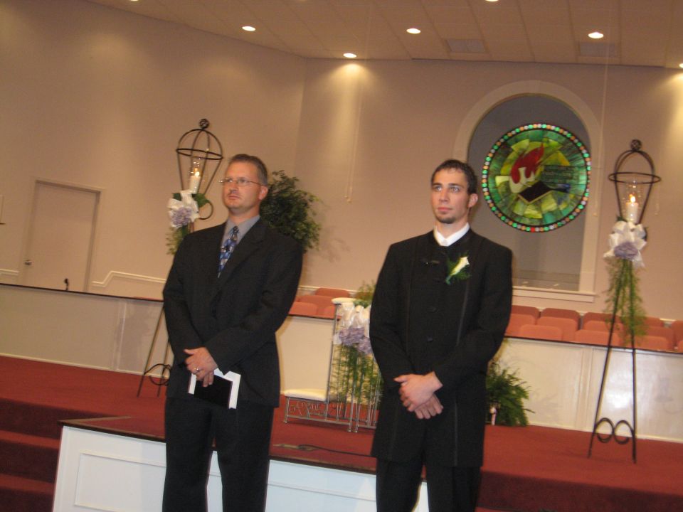 Two men in formal wear stand on a red carpet in a church, with decorative lamps and stained glass visible