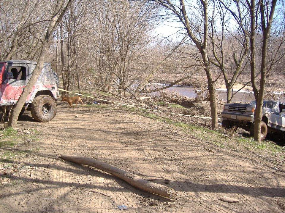 Red and grey Jeep Wrangler with large tires, 3/4 front view, parked on a dirt path next to a river