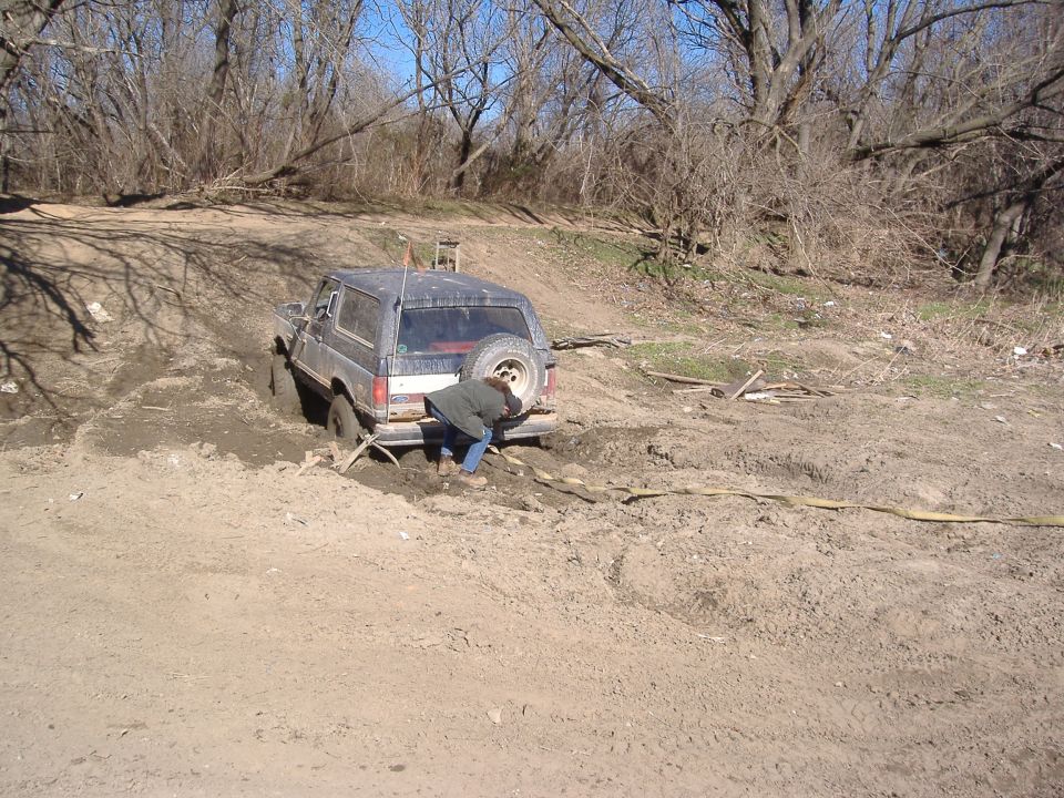Dark Ford Bronco stuck in mud, spare tire on rear door, person pushing