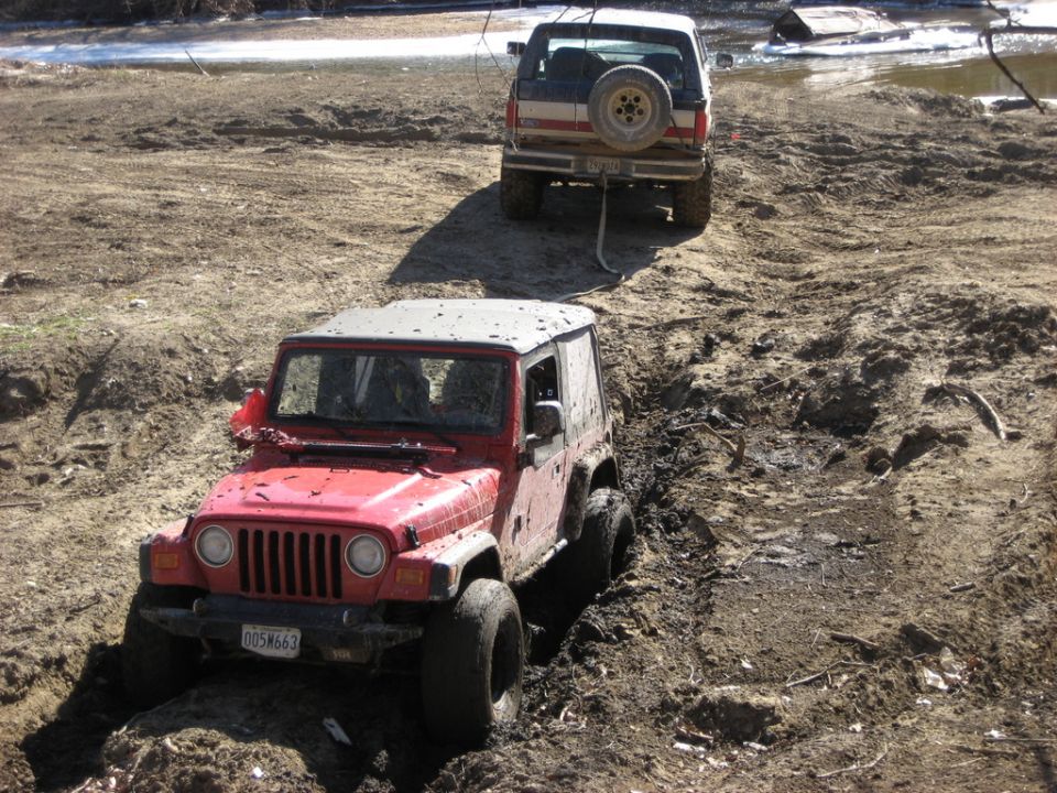 Muddy red Jeep Wrangler stuck in a mud pit, strapped to a lifted red and white Bronco