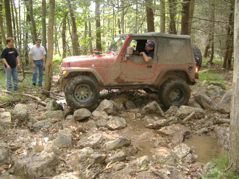 2003 Jeep Wrangler navigating a rocky, muddy trail with two people standing nearby