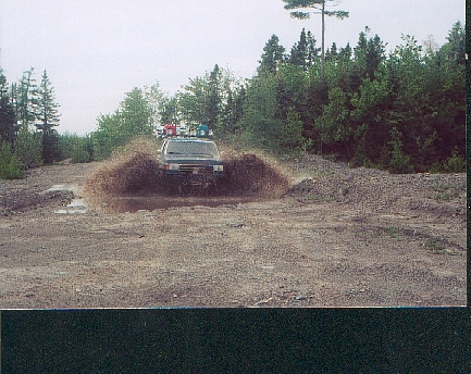1989 Ford Bronco - An off-road vehicle powers through a muddy obstacle, creating a large splash.