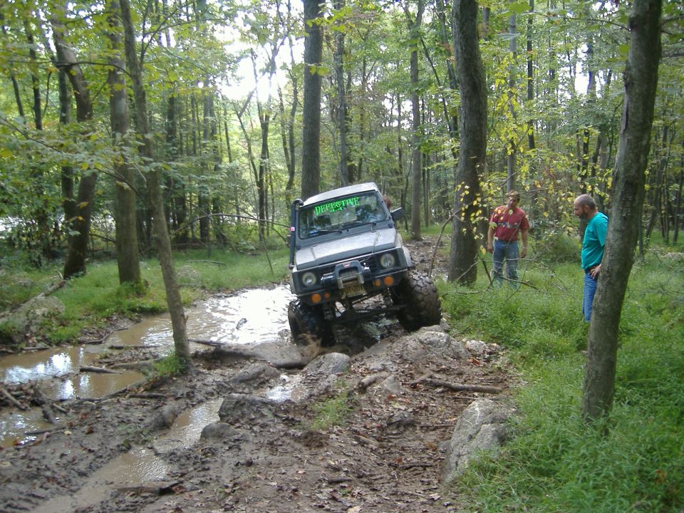 Suzuki Samurai navigating a muddy trail with large tires and a winch. Two people watch from the side