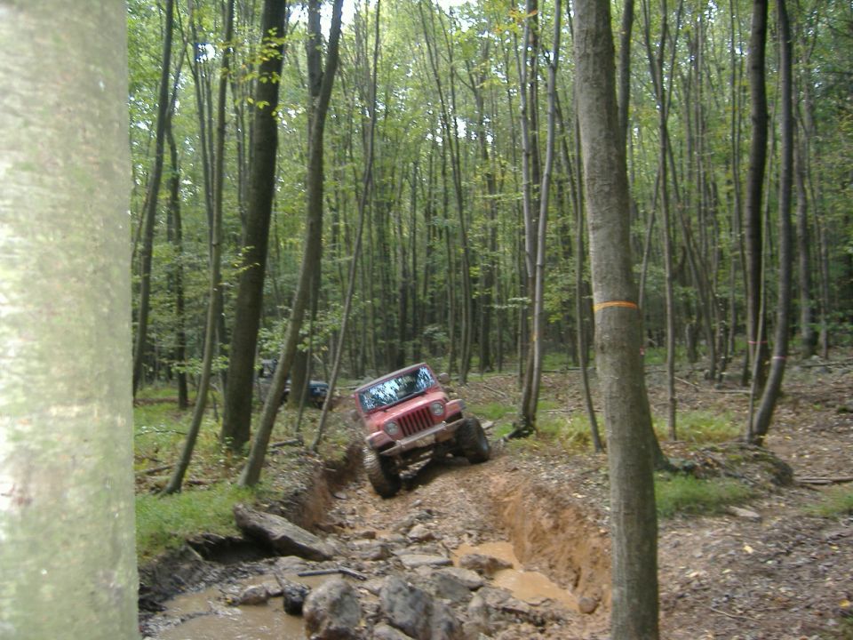Red Jeep Wrangler driving through a muddy, wooded trail. Another Jeep is visible in the background