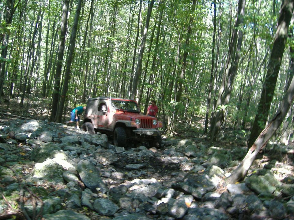 Red 2003 Jeep Wrangler navigating a rocky forest trail with two people nearby