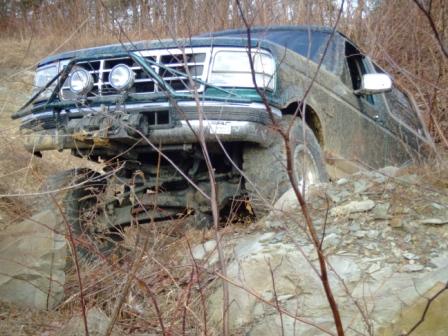 1996 Ford Bronco - A dark green lifted Ford Bronco crawls over a rock ledge on a wooded trail, front driver-side wheel drooped into a hole with the suspension…