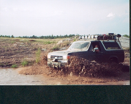 Ford Bronco driving through mud, kicking up water and dirt. It has a roof rack with gear