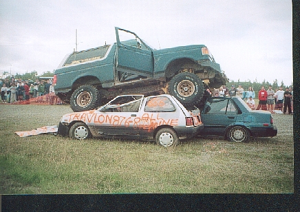 1989 Ford Bronco - A large Ford Bronco is crushing smaller cars in a grassy field with spectators in the background.