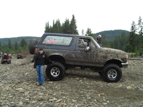1995 Ford Bronco - A muddy Ford Bronco is parked on a rocky trail with ATVs in the distance. A woman stands next to the vehicle.