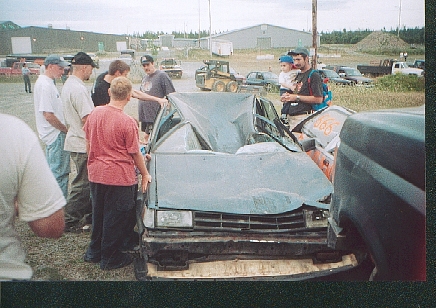 1989 Ford Bronco - A group of people gather around a severely damaged sedan in an outdoor lot.