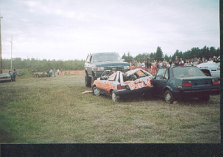 1989 Ford Bronco - A 1989 Ford Bronco looms over a heavily damaged car, likely from a demolition derby event.