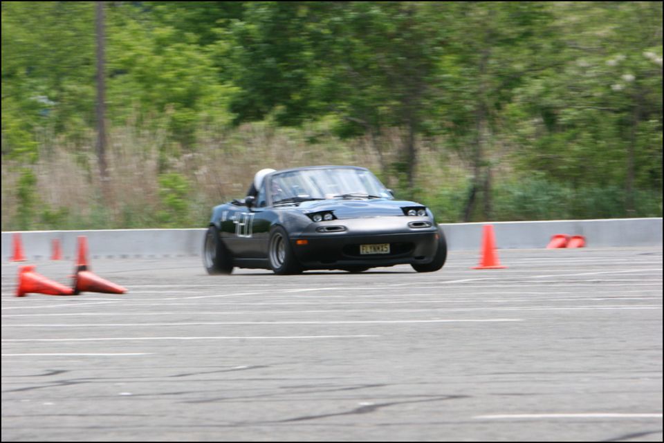 Black Mazda Miata MX-5 with racing numbers on door, driving on a track with orange cones