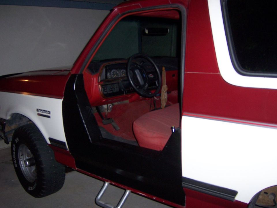 Red and white Ford Bronco with driver door open showing red interior, parked in garage