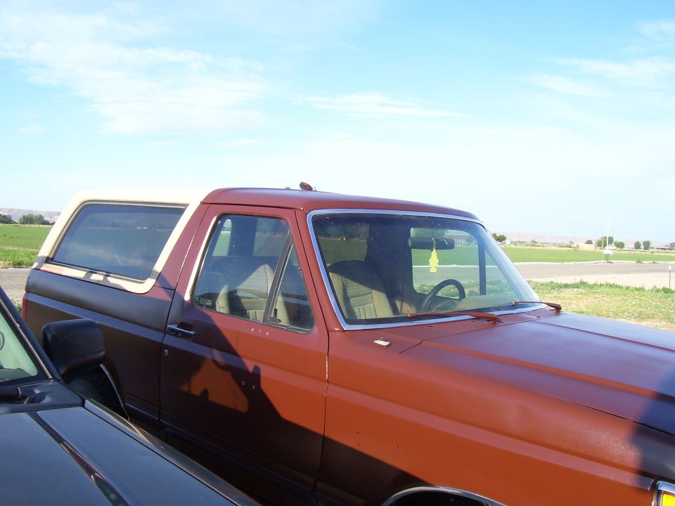 Side view of a brown Ford Bronco with a white top. Another dark vehicle is in the foreground