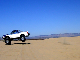 1996 Ford Bronco - A white and blue off-road truck is captured airborne over a sandy desert landscape.