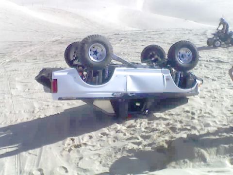 1996 Ford Bronco - An overturned white SUV rests on a sandy terrain, with a person on an ATV visible in the distance.