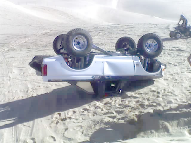 White SUV overturned on sand dunes, showing its underside and wheels. A person rides an ATV in the background