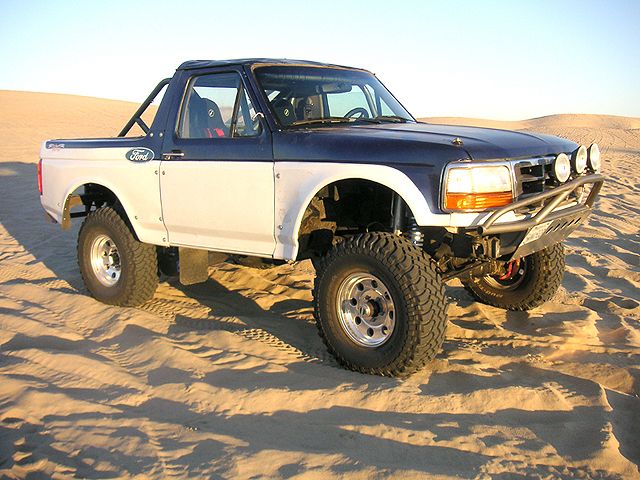Topless two-tone Ford Bronco prerunner with cage and tube bumper parked on sand dunes