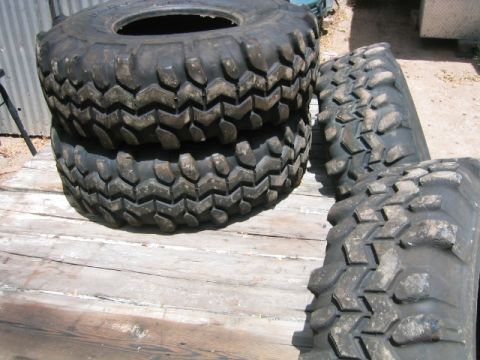 1973 Ford F-100 - Four unmounted mud-terrain tires with aggressive chunky lug tread, two stacked flat and two standing on edge, on a weathered wood deck besi…