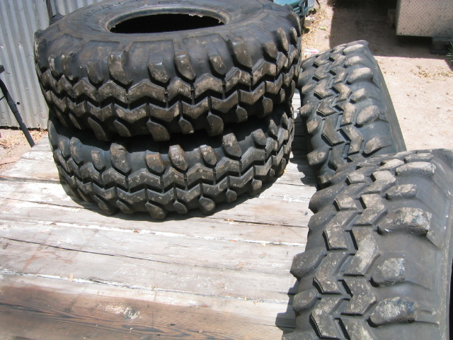 Four used mud-terrain tires with chunky lug tread stacked on a wood deck