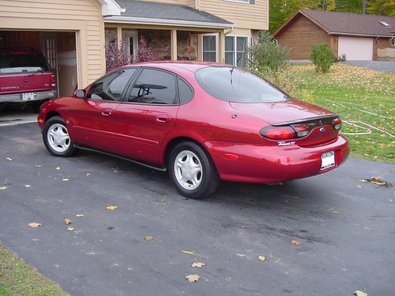 Dark red four-door sedan, rear three-quarter view, parked on driveway by tan garage
