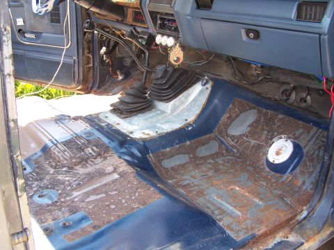 1986 Ford Bronco - The interior floor of a vehicle is exposed, revealing rust and exposed wiring, with dual gear shifters in place.