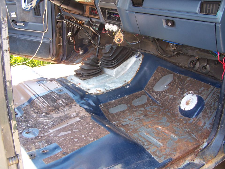 Interior of a vehicle with floor removed, showing rust and wiring. Two gear shifters are visible