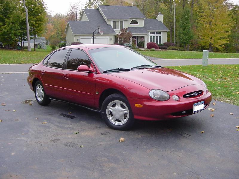 Dark red Ford Taurus sedan parked on a leaf-covered driveway, front passenger-side view, house behind