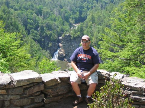 1985 Ford LTD Crown Victoria - A man wearing a NASCAR shirt and shorts sits on a stone wall, with a waterfall and lush green trees in the background.