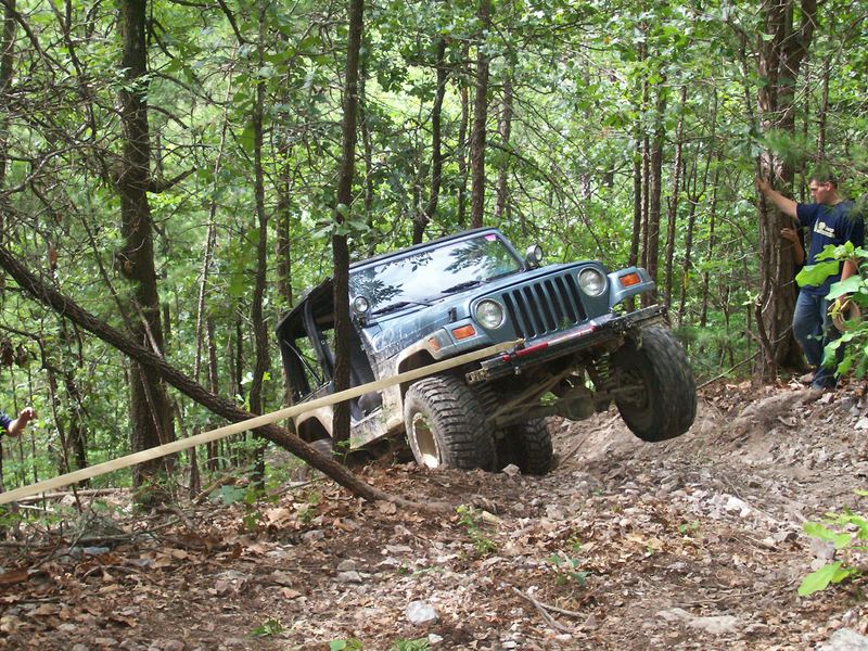 Blue Jeep Wrangler on a steep, wooded trail, with one front wheel lifted