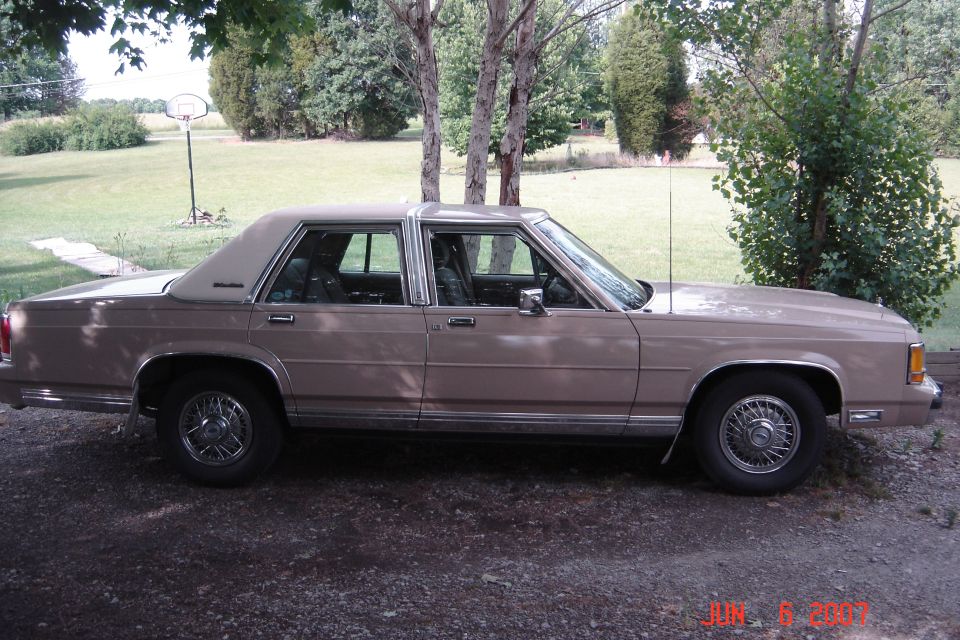 Tan 1989 Ford LTD Crown Victoria sedan, passenger side view, parked on gravel by a grassy yard